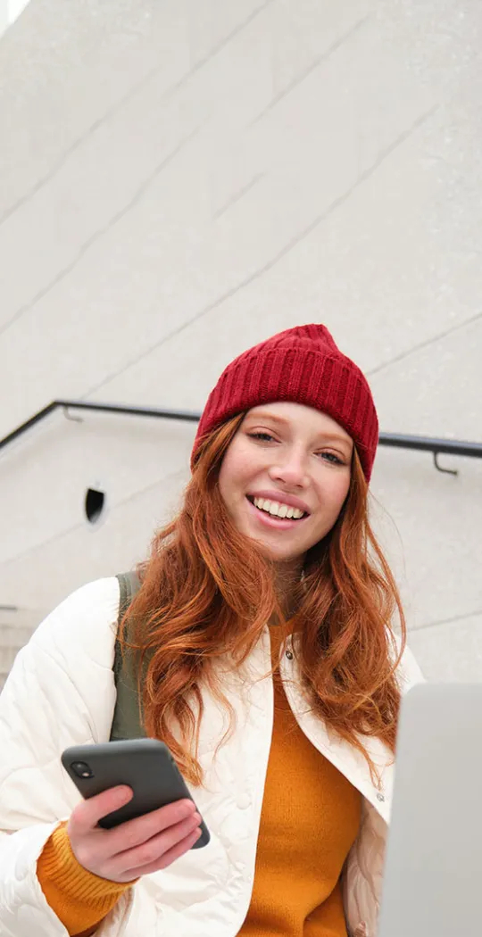 Junge Frau sitzt mit Smartphone und Laptop auf einer Treppe. 